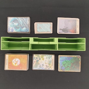 May include: A green plastic card organizer with multiple compartments, holding several stacks of illustrated cards. The cards feature various fantasy art, including landscapes and abstract designs. The organizer is on a black surface.