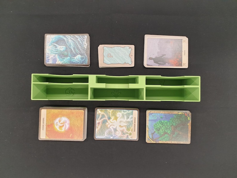 May include: A green plastic card organizer with multiple compartments, holding several stacks of illustrated cards. The cards feature various fantasy art, including landscapes and abstract designs. The organizer is on a black surface.