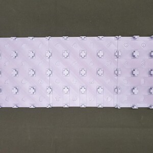 May include: A light purple interlocking tile set, made up of several rectangular pieces. Each tile features a grid of cross-shaped connectors and small, embossed shapes. The tiles are designed to interlock, forming a larger surface area.