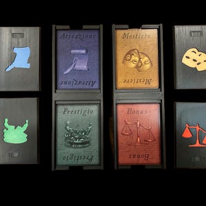 May include: Eight black wooden tiles with different colored symbols and text. The tiles are arranged in two rows of four. The top row has a blue symbol, a purple tile with the text "Attrazione", a gold tile with the text "Mestiere", and a tile with two yellow masks. The bottom row has a green symbol, a green tile with the text "Prestigio", a red tile with the text "Bonus", and a tile with a red scale.