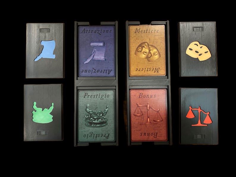 May include: Eight black wooden tiles with different colored symbols and text. The tiles are arranged in two rows of four. The top row has a blue symbol, a purple tile with the text "Attrazione", a gold tile with the text "Mestiere", and a tile with two yellow masks. The bottom row has a green symbol, a green tile with the text "Prestigio", a red tile with the text "Bonus", and a tile with a red scale.
