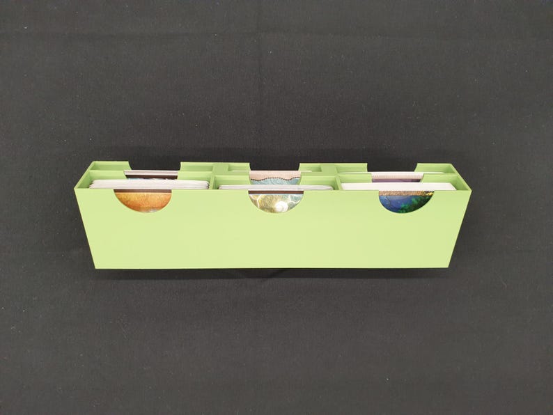 May include: A light green, rectangular card organizer with three curved cutouts. The organizer is divided into four sections, each holding a stack of colorful cards. The cards have various designs and colors.