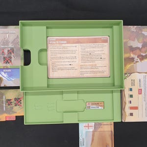 May include: An open, light green board game box with various game components. The box contains cards with text, a card holder, and other game elements. The game cards have text in Italian. The game is set against a dark background.