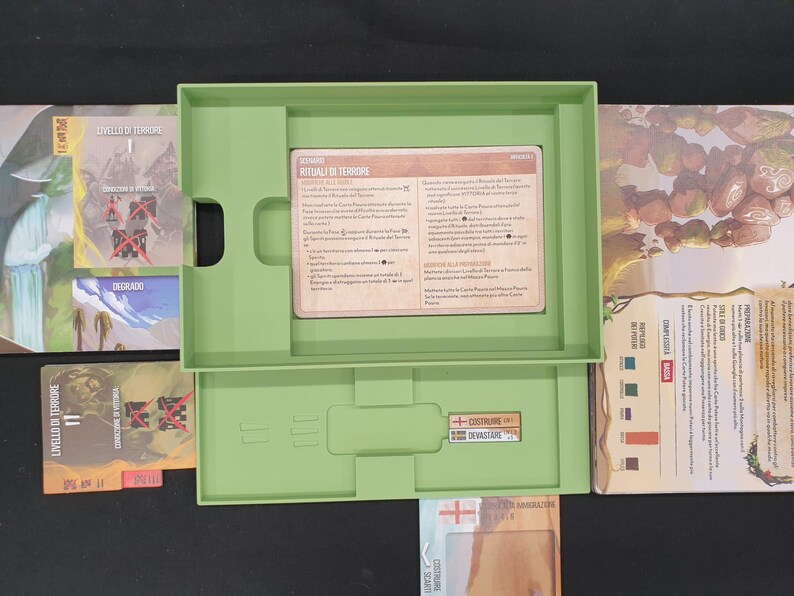 May include: An open, light green board game box with various game components. The box contains cards with text, a card holder, and other game elements. The game cards have text in Italian. The game is set against a dark background.