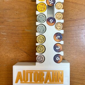 Pode incluir: Organizador de pl&aacute;stico branco para o jogo de tabuleiro Autobahn. O organizador tem slots para diferentes fichas coloridas, incluindo 1, 2, 5, 9, 10 e 20. O organizador tem a palavra "AUTOBAHN" impressa na frente em letras douradas.