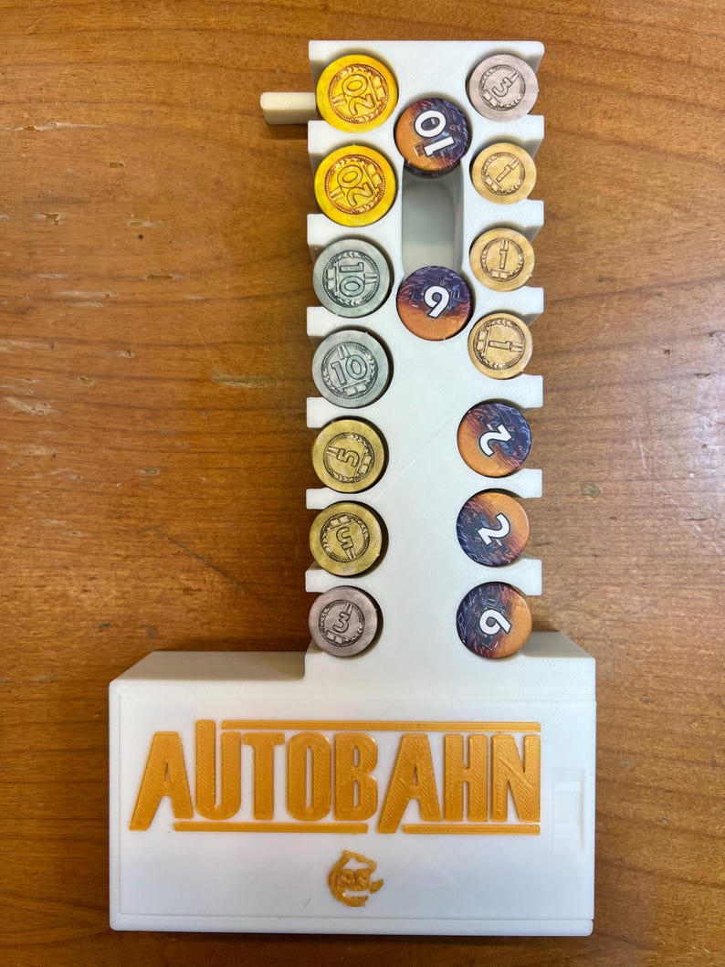 Pode incluir: Organizador de pl&aacute;stico branco para o jogo de tabuleiro Autobahn. O organizador tem slots para diferentes fichas coloridas, incluindo 1, 2, 5, 9, 10 e 20. O organizador tem a palavra "AUTOBAHN" impressa na frente em letras douradas.