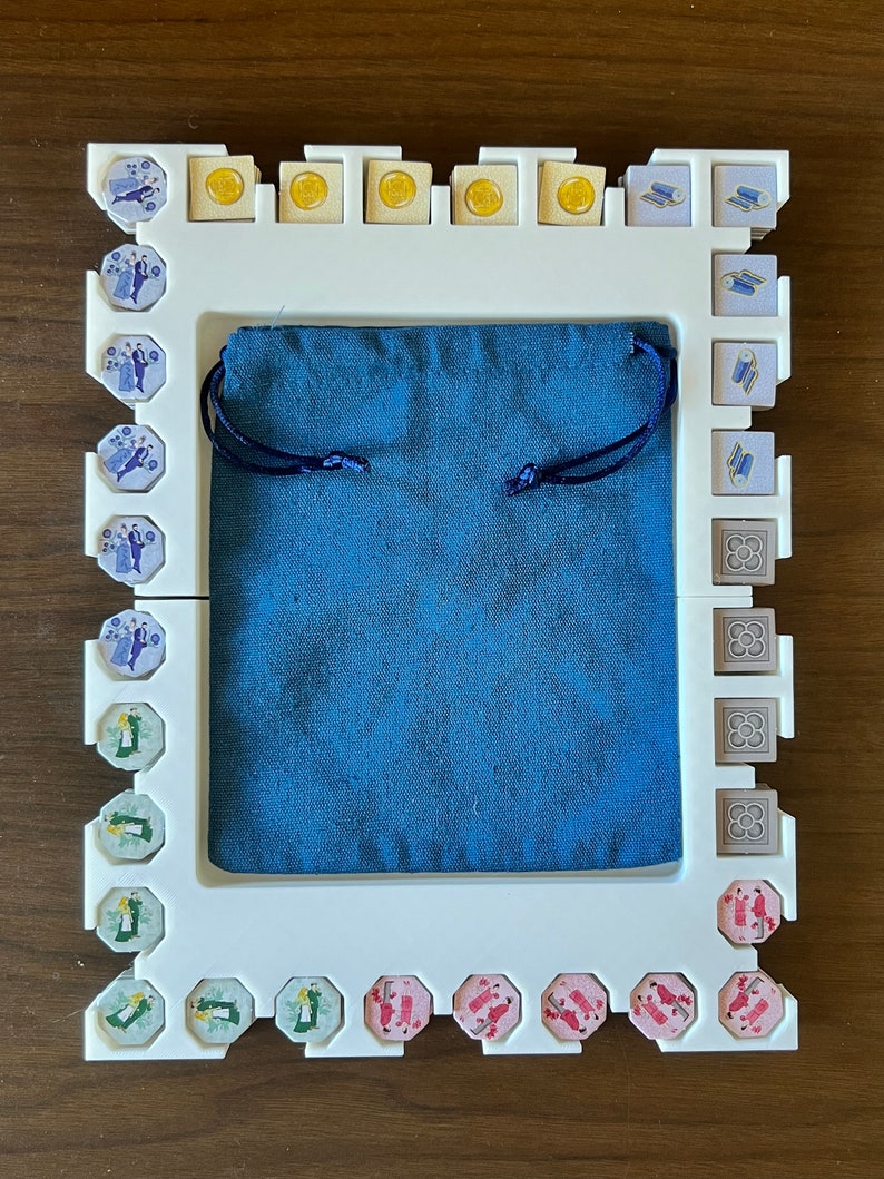 K&ouml;nnte beinhalten: Ein wei&szlig;er Rahmen mit einem blauen Stoffbeutel im Inneren. Der Rahmen hat einen Rand aus kleinen, bunten Fliesen mit verschiedenen Designs. Die Fliesen sind in einem Muster um den Beutel angeordnet.