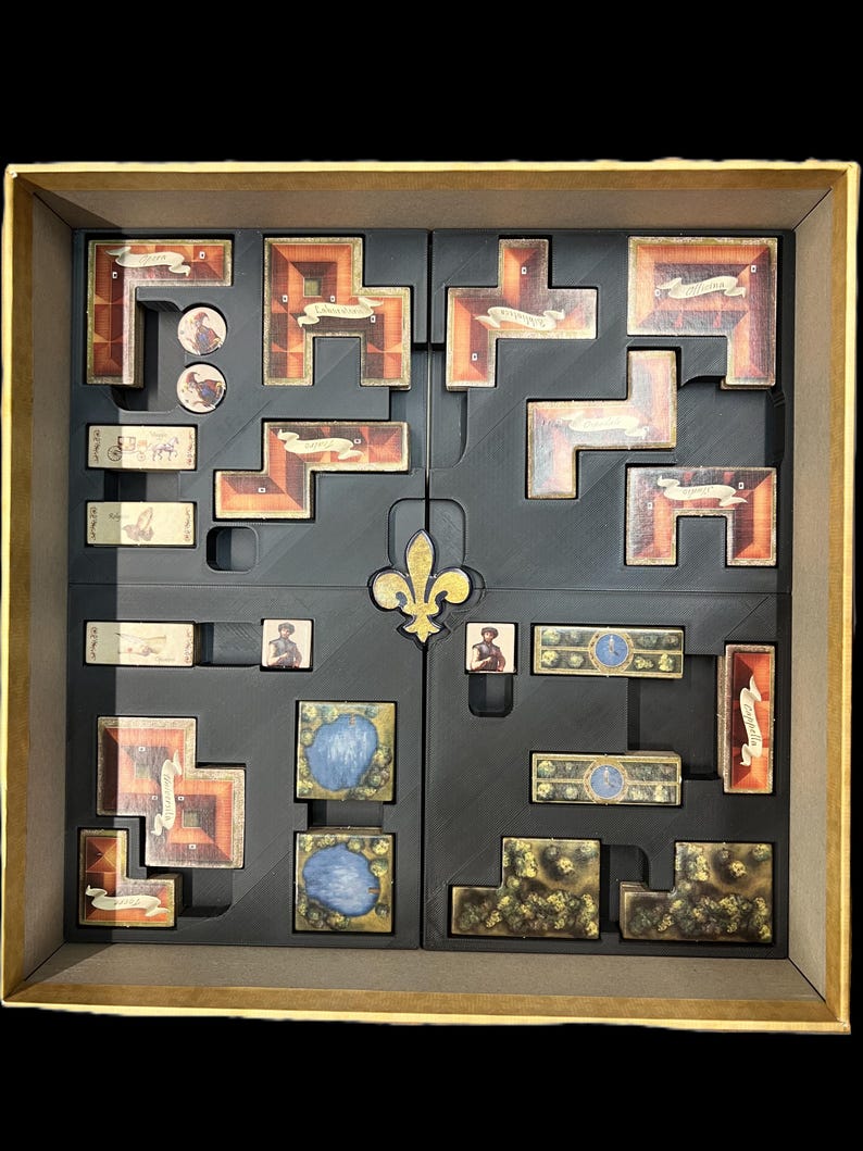 K&ouml;nnte beinhalten: Ein schwarzer Schaumstoffeinsatz mit F&auml;chern zur Aufbewahrung von Spielsteinen. Der Einsatz hat in der Mitte ein Fleur-de-lis-Design und F&auml;cher zur Aufbewahrung verschiedener Spielsteine, darunter Kacheln, Karten und Figuren.