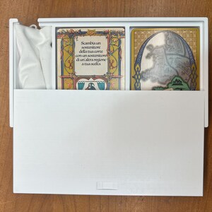 Op de afbeelding: Een witte doos met een witte doek erin. De doos bevat twee kaarten. De kaart links heeft een gouden lijst met een kleurrijk ontwerp en tekst in het Italiaans. De kaart rechts heeft een gouden lijst met een kaart erin.