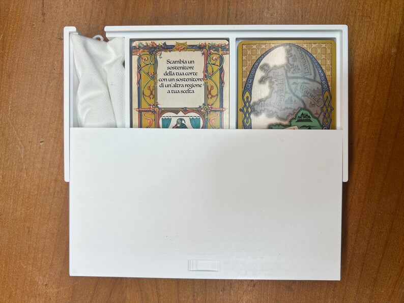 Op de afbeelding: Een witte doos met een witte doek erin. De doos bevat twee kaarten. De kaart links heeft een gouden lijst met een kleurrijk ontwerp en tekst in het Italiaans. De kaart rechts heeft een gouden lijst met een kaart erin.