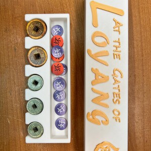 May include: A white game board with a compartment for storing game pieces. The board has the words "At the Gates of Loyang" printed on it in gold lettering. The board is next to a white tray with compartments for storing game pieces. The tray has a variety of game pieces in it, including coins and tokens.