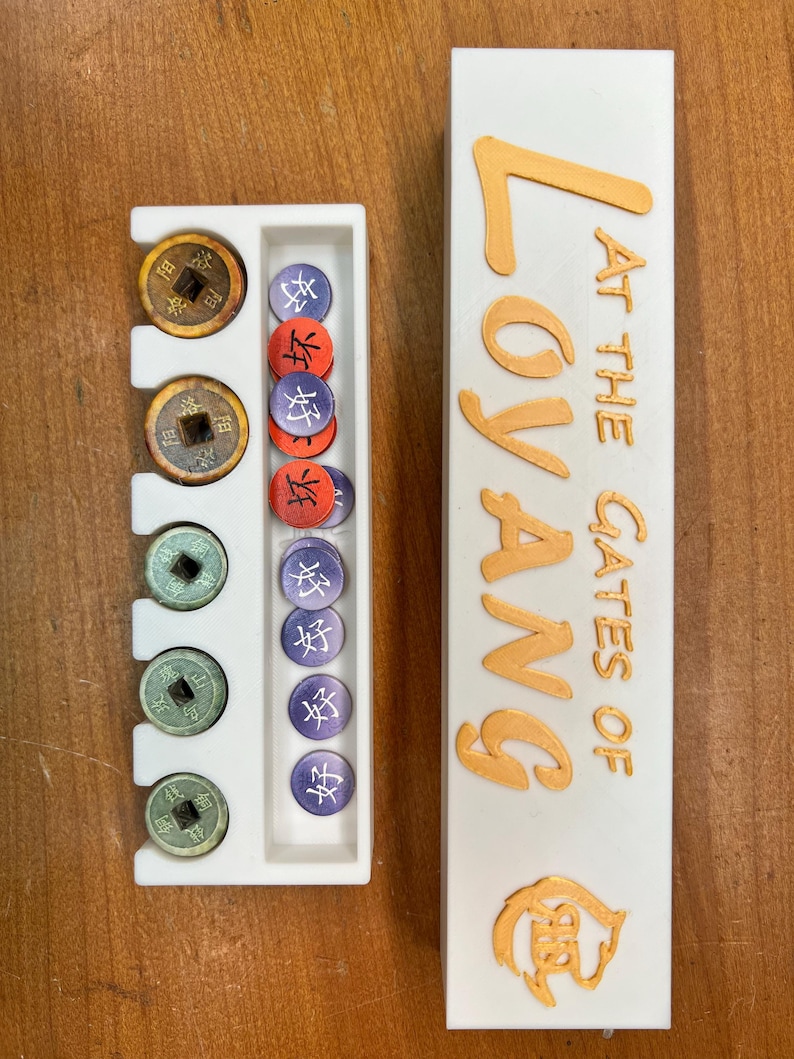 May include: A white game board with a compartment for storing game pieces. The board has the words "At the Gates of Loyang" printed on it in gold lettering. The board is next to a white tray with compartments for storing game pieces. The tray has a variety of game pieces in it, including coins and tokens.