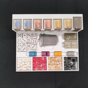 May include: A white board game organizer filled with game components. Includes dice, small wooden cubes in various colors, game cards, and cylindrical pieces. The organizer is on a black surface.