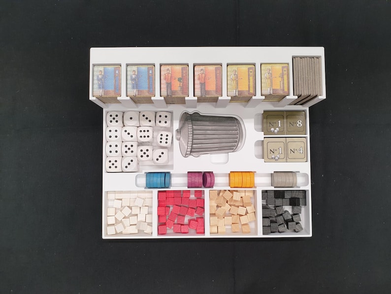 May include: A white board game organizer filled with game components. Includes dice, small wooden cubes in various colors, game cards, and cylindrical pieces. The organizer is on a black surface.