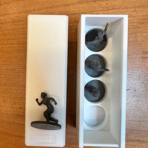 May include: A white rectangular container with a running figure figurine on a brown base. Next to it is a white container with three running figure figurines in circular compartments and a white sphere.