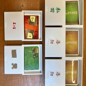 May include: Five white boxes with lids open, revealing game cards. The boxes are labeled with Chinese characters and the word "LOYANG". The cards feature illustrations of buildings, animals, and numbers.