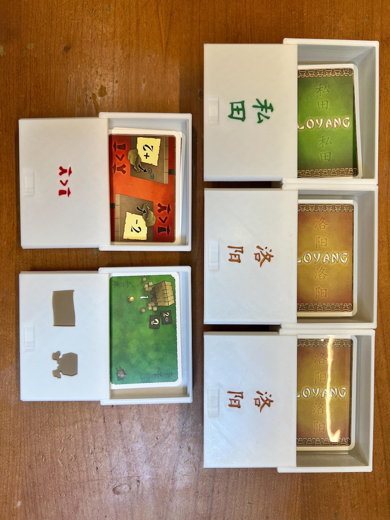 May include: Five white boxes with lids open, revealing game cards. The boxes are labeled with Chinese characters and the word "LOYANG". The cards feature illustrations of buildings, animals, and numbers.