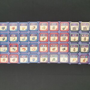 May include: A selection of game tiles arranged in rows. The tiles are square with various icons and colours, including blue, red, and purple. The icons depict items like food, tools, and resources. The tiles are set in a purple plastic frame.