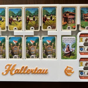 May include: A white plastic game organizer with the word "Hallertau" printed in gold. The organizer has slots for cards and a section for tokens. The cards feature illustrations of farm scenes and gates. The tokens are for a bakery and have a wheat symbol and a number 1.