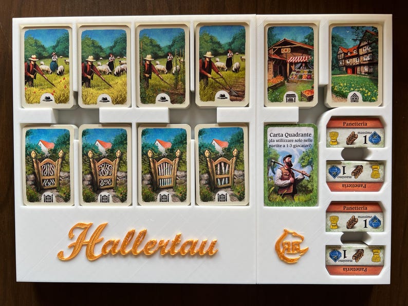 May include: A white plastic game organizer with the word "Hallertau" printed in gold. The organizer has slots for cards and a section for tokens. The cards feature illustrations of farm scenes and gates. The tokens are for a bakery and have a wheat symbol and a number 1.