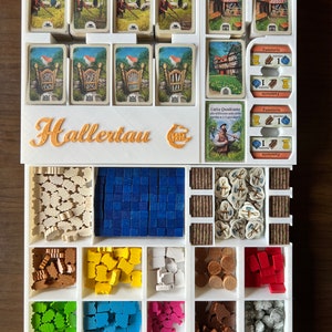 May include: A white plastic organizer with compartments for storing game pieces and cards. The organizer has the words "Hallertau" printed on the front. The organizer is filled with various game pieces, including wooden tiles, meeples, and cards.