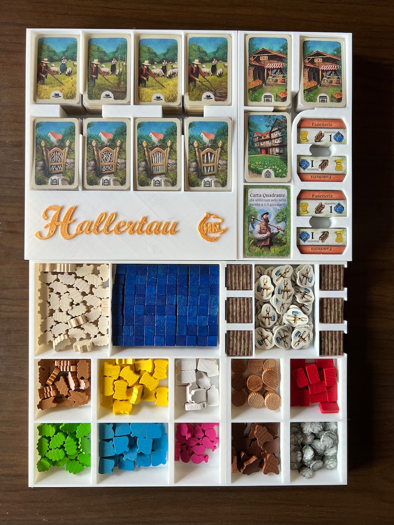 May include: A white plastic organizer with compartments for storing game pieces and cards. The organizer has the words "Hallertau" printed on the front. The organizer is filled with various game pieces, including wooden tiles, meeples, and cards.
