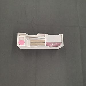 May include: A white plastic card organiser with several compartments. It contains stacks of cards, a pink circular piece, and other small items. The organiser is designed to keep cards and accessories neatly arranged.