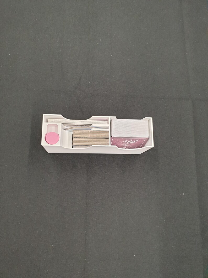 May include: A white plastic card organiser with several compartments. It contains stacks of cards, a pink circular piece, and other small items. The organiser is designed to keep cards and accessories neatly arranged.