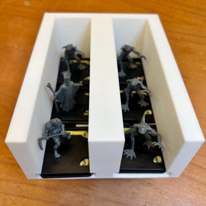 May include: A white, rectangular game organizer with two compartments. Each compartment holds several small, dark-colored cards with yellow markings and miniature gray figurines. The figurines depict various fantasy creatures and characters.