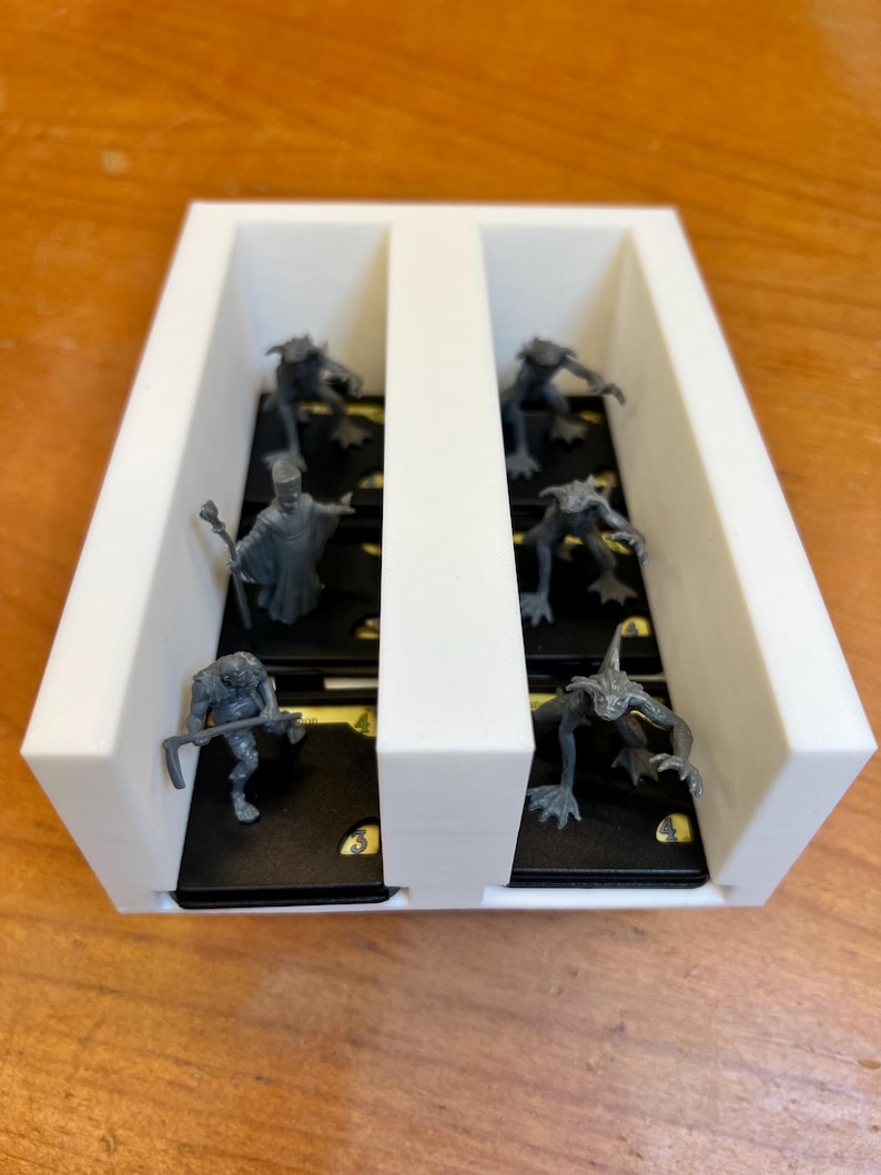 May include: A white, rectangular game organizer with two compartments. Each compartment holds several small, dark-colored cards with yellow markings and miniature gray figurines. The figurines depict various fantasy creatures and characters.