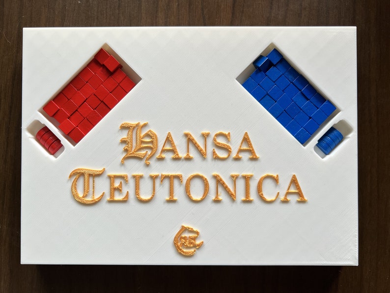 Może przedstawiać: Białe pudełko gry planszowej z tytułem "HANSA TEUTONICA" złotymi literami. Pudełko ma dwa przedziały, jeden z czerwonymi kostkami, a drugi z niebieskimi kostkami.