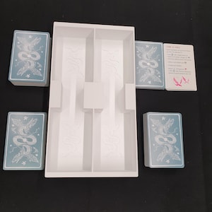 May include: A white game organiser with several compartments, surrounded by card decks. The cards feature a light blue background with a detailed illustration of a serpent and floral elements. A card with text is also visible.