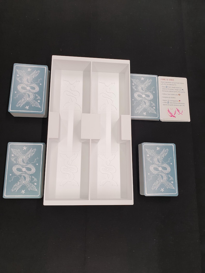 May include: A white game organiser with several compartments, surrounded by card decks. The cards feature a light blue background with a detailed illustration of a serpent and floral elements. A card with text is also visible.
