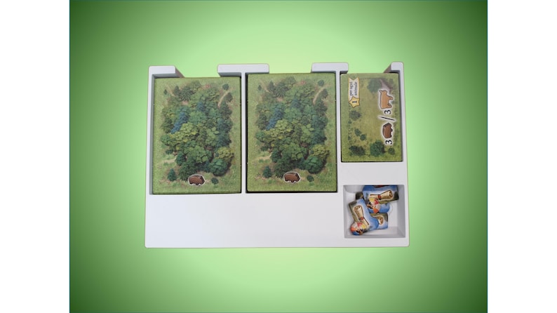 May include: White plastic organizer with three compartments for storing game pieces. The compartments contain green and brown illustrated cards and small blue and white game pieces.