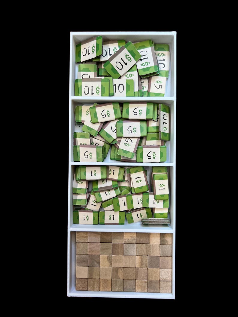 May include: A white box with three compartments containing stacks of green and brown game pieces. The top compartment contains $10 bills, the middle compartment contains $5 bills, and the bottom compartment contains $1 bills. The bottom compartment also contains a stack of small wooden blocks.
