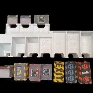 May include: A white plastic game organizer with labeled compartments for cards and game pieces. The organizer has slots labeled "GREEN", "BLUE", "EXIT", "RED", and others. Several stacks of cards and game tokens are visible.