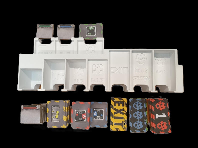 May include: A white plastic game organizer with labeled compartments for cards and game pieces. The organizer has slots labeled "GREEN", "BLUE", "EXIT", "RED", and others. Several stacks of cards and game tokens are visible.
