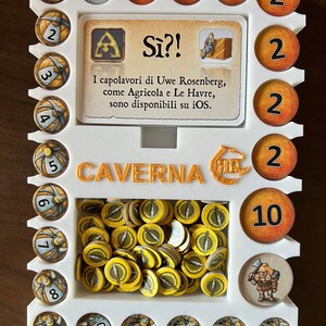 May include: A white plastic game organizer with the words "CAVERNA" and "S&igrave;?!" printed on it. The organizer has slots for different game pieces, including wooden tokens, a card, and a small figure. The organizer is designed for the board game "Caverna: The Cave Farmers".