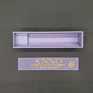 May include: A lavender-coloured rectangular box with a divider and a separate lavender-coloured rectangular piece. The piece has the text "ANNO 1800" in gold lettering, along with a gold emblem.