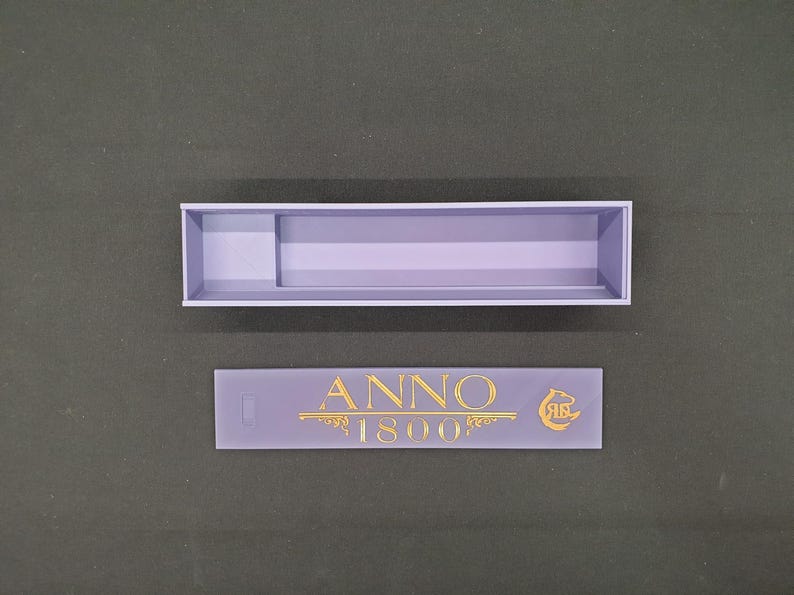 May include: A lavender-coloured rectangular box with a divider and a separate lavender-coloured rectangular piece. The piece has the text "ANNO 1800" in gold lettering, along with a gold emblem.