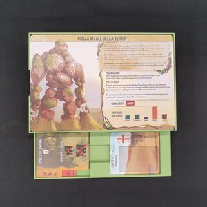 May include: A green box containing game cards with illustrations and text. The top card features a stone golem illustration and the text "FORZA VITALE DELLA TERRA". Other cards show various graphics and text, with a black background.
