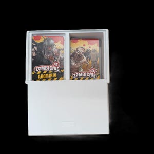 May include: A white box containing two decks of Zombicide game cards. The cards feature zombie illustrations and the game title. The box is open, revealing the card decks inside. The background is black.