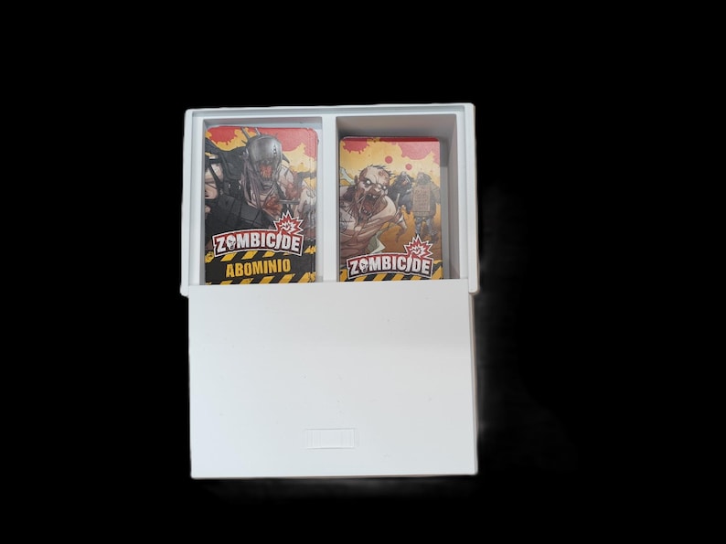 May include: A white box containing two decks of Zombicide game cards. The cards feature zombie illustrations and the game title. The box is open, revealing the card decks inside. The background is black.