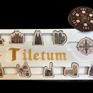 May include: A white game board with the word "Tiletum" printed in gold letters. The board has nine slots for game pieces, each with a number from 1 to 9. There are also two additional slots for a compass and a die. The game pieces are small, detailed images of buildings.