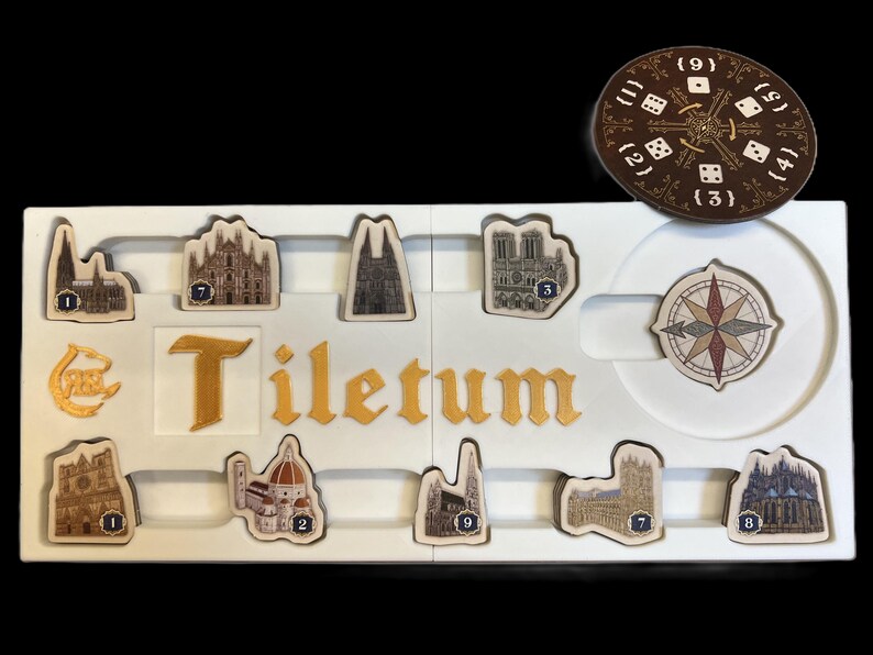 May include: A white game board with the word "Tiletum" printed in gold letters. The board has nine slots for game pieces, each with a number from 1 to 9. There are also two additional slots for a compass and a die. The game pieces are small, detailed images of buildings.