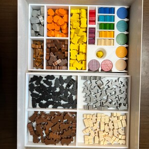 May include: A white box with a white insert containing various wooden game pieces. The pieces are organized by color and shape, including gray, orange, yellow, brown, black, and white. The box is open and the insert is visible.