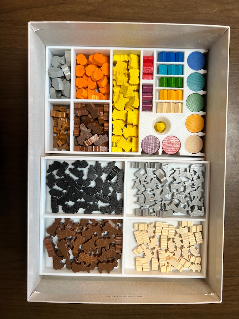 May include: A white box with a white insert containing various wooden game pieces. The pieces are organized by color and shape, including gray, orange, yellow, brown, black, and white. The box is open and the insert is visible.