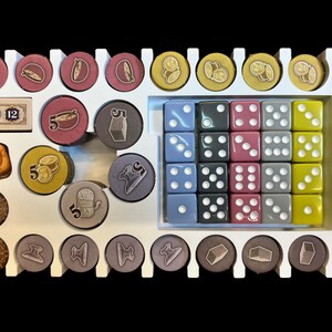 May include: A white organizer with compartments for storing game pieces. The organizer contains various colored dice, small wooden tokens, and small round pieces with numbers and images. The organizer has the numbers 6 and 12 printed on it.