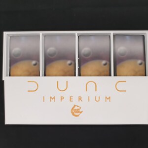 May include: A white box containing four game card stacks with a planetary design. The box has the words "DUNE IMPERIUM" in orange lettering, along with a graphic. The background is black.
