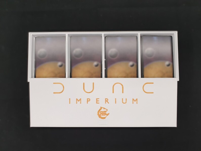 May include: A white box containing four game card stacks with a planetary design. The box has the words "DUNE IMPERIUM" in orange lettering, along with a graphic. The background is black.